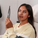 Mujer joven de pelo largo oscuro sentada en un sillón blanco observa su nariz en un espejo de mano, detrás una persona con guantes negros.