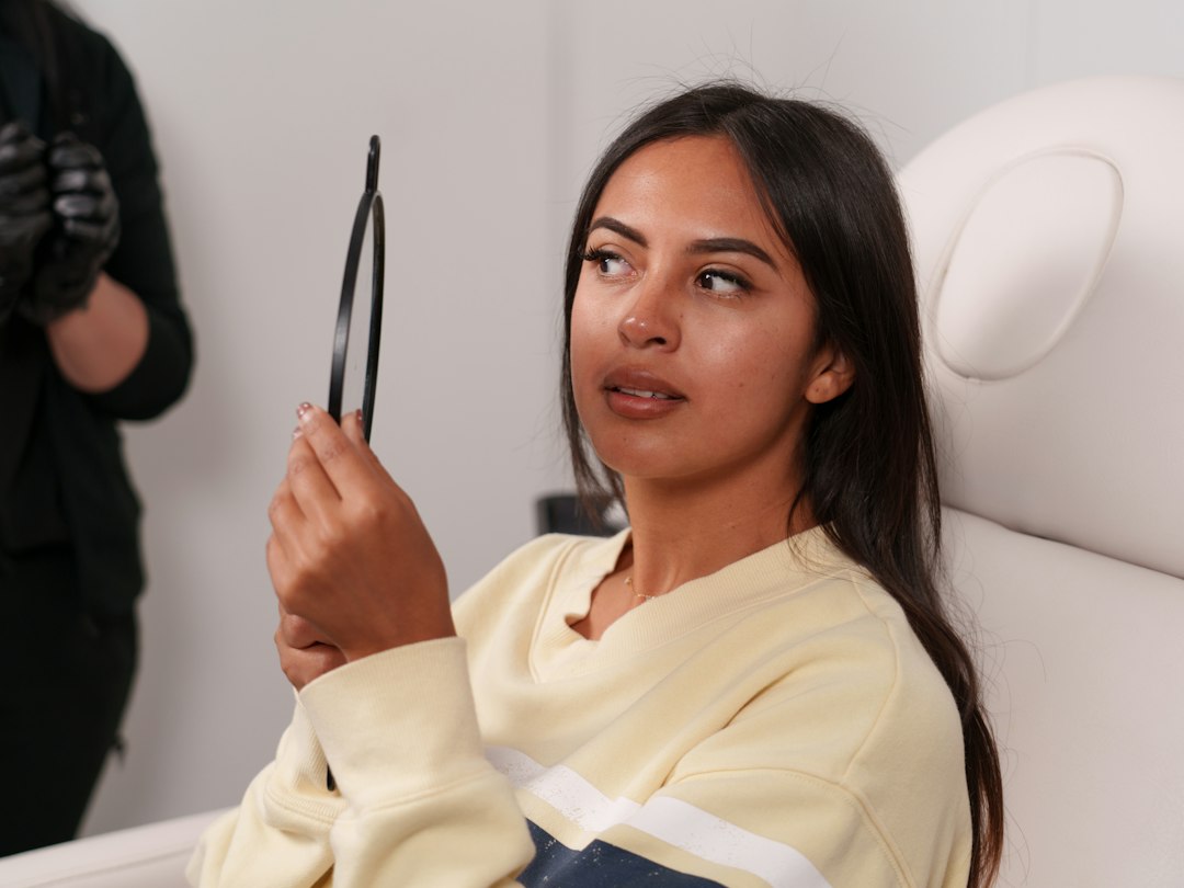 Mujer joven de pelo largo oscuro sentada en un sillón blanco observa su nariz en un espejo de mano, detrás una persona con guantes negros.